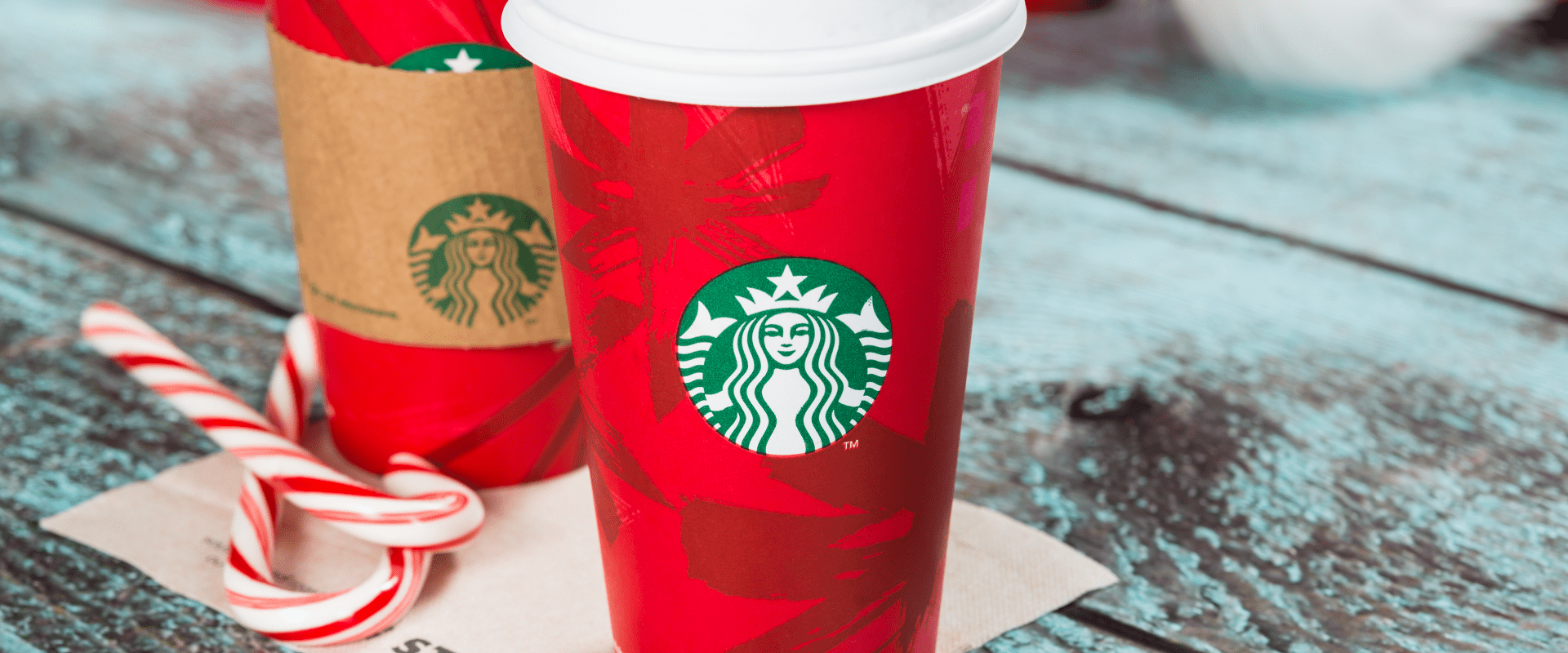 Red Starbucks holiday cups with the green siren logo sit on a rustic blue tabletop with peppermint candy canes in the foreground, featured for Lake Crest Village’s holiday season.